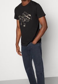 Svart t-shirt i bomull med ett tekniskt diagram tryckt i beige. Korta ärmar och rund halsringning. Matchas med mörkblå jeans.