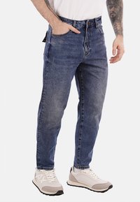 Jeans in denim blu con un design a gamba dritta, cinque tasche e passanti per cintura. Indossati con scarpe da ginnastica di colore chiaro con dettagli grigi.