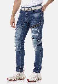 Blaue, stark ausgebleichte Skinny Jeans mit Rissen und gepatchten Details, versehen mit einem schwarzen Logosstreifen, kombiniert mit einem weißen Nieten-Gürtel und weißen Sneakers.