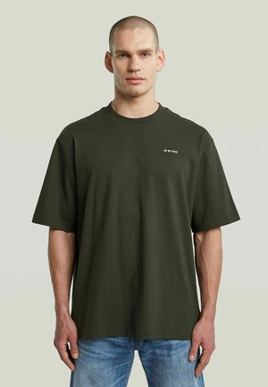 Man in een donkergroene G-Star oversized t-shirt en blauwe jeans, staand tegen een effen lichte achtergrond.