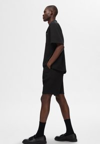 Fekete bordás póló és nadrág, mellzsebbel és laza szabással. Fekete zoknival és cipővel viselve, oldalnézet világos háttér előtt.