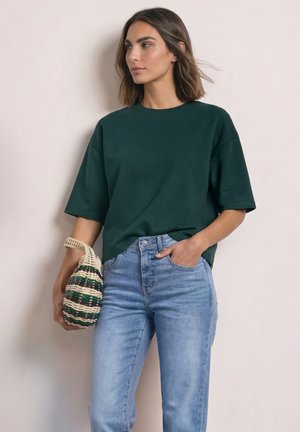Kvinde iført en mørkegrøn oversized T-shirt og lyseblå jeans, der holder en vævet stribet håndtaske og står mod en ensfarvet baggrund.