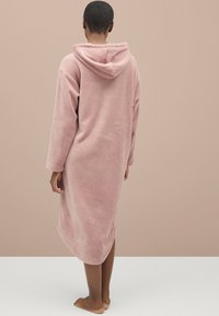Lyserød hættebadekåbe lavet af blødt fleece-materiale. Har lange ærmer og en let buet nederste kant. Båret af en person set fra ryggen.
