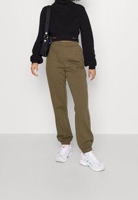 Czarny ryfrowany sweter z wysokim kołnierzem o skróconym kroju, zestawiony z oliwkowymi spodniami dresowymi z haftowanym logo. Białe sneakersy dopełniają look.