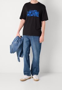 Persona que lleva una camiseta gráfica negra con el texto azul "HUGO", jeans azules, zapatillas blancas, sosteniendo una chaqueta de mezclilla azul en la mano izquierda.