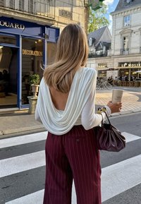 Weißes Oberteil mit Drapierung und offenem Rücken, kombiniert mit bordeauxroten, gestreiften weit geschnittenen Hosen. Hält eine strukturierte Tasse und eine dunkelbraune Handtasche.