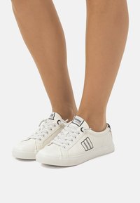 Zapatillas blancas con acentos y logotipo en negro. Material de cuero suave, puntera redonda, suela plana y diseño de cordones. Construcción duradera.