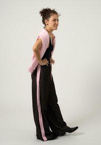 Donna con capelli ricci raccolti, che indossa un top nero senza maniche, pantaloni larghi stampa leopardata con strisce rosa e un maglione rosa appoggiato sulle spalle.