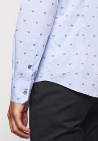 Jasnoniebieska koszula z długim rękawem z drobnym, powtarzającym się ciemnym wzorem logo, zapiętymi mankietami, noszona z czarnymi spodniami, prezentująca lewą rękę i widok z boku.