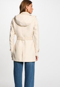 Manteau ajusté beige avec une capuche, taille ceinturée et tissu texturé. Comprend des poignets boutonnés et une fente au dos pour plus de liberté de mouvement.