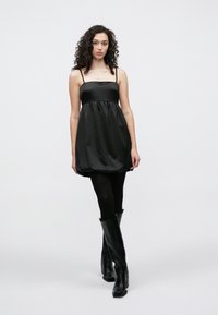 Μίνι μαύρο σατέν φόρεμα με λεπτά τιράντες και σφηκωτή μέση, με φούστα σε σιλουέτα φούσκας. Φορεμένο με μαύρες μπότες μέχρι το γόνατο.