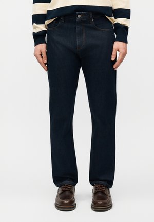 Bărbat purtând blugi bleu închis cu picior drept, pantofi maro din piele și un pulover bej cu dungi bleumarin, stând în picioare pe un fundal simplu.