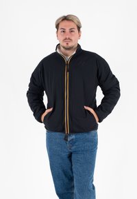 Czarna kurtka zapinana na zamek błyskawiczny z dwiema przednimi kieszeniami, z żółtym zamkiem i logo na górnej lewej piersi. Noszona z niebieskimi dżinsami.