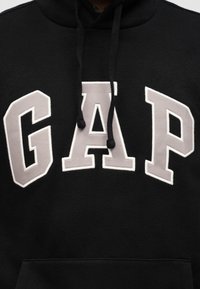 Μαύρο φούτερ με κουκούλα με μεγάλα γκρι γράμματα με λευκή περίγραμμα που σχηματίζουν τη λέξη "GAP" στο στήθος και μαύρα κορδόνια που κρέμονται μπροστά.