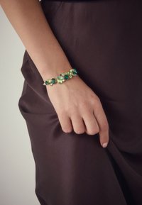 Green gemstone bracelet featuring various shapes and sizes, set in gold-toned hardware, worn on a hand against a dark fabric background.