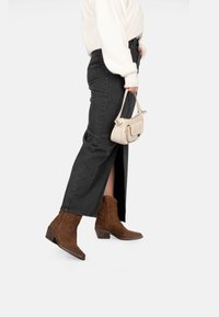 Stivali cowboy alla caviglia in suede marrone con punta affusolata e dettagli ricamati, abbinati a una gonna nera maxi e un maglione color crema.