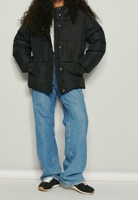 Persona vistiendo una chaqueta acolchada negra, jeans de pierna ancha azul claro y zapatillas negras y blancas, de pie contra una pared clara y lisas.