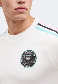 Бяла суитшърт на млад мъж с лого на Club Internacional de Fútbol Miami MMXX и розови, черни и тюркоазени ивици на рамото.