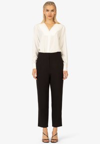 Weiße Langarmbluse mit V-Ausschnitt, kombiniert mit hochgeschnittenen schwarzen Hosen und schwarzen Riemchenheels. Verfügt über klare Linien und minimalistisches Design.