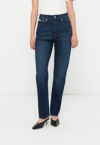 Jean en denim bleu foncé avec une coupe droite, taille haute, poches avant et quincaillerie argentée. Associé à des chaussures à talons noires.