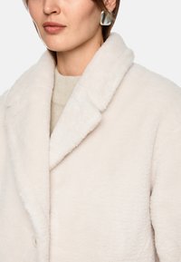 Hellbeige Oversized-Mantel mit weicher, flauschiger Textur, Reverskragen und Knopfverschluss, getragen über einem leichten Pullover. Silbern geometrische Ohrringe setzen Akzente.