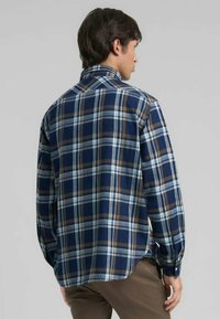 Hombre de perfil vistiendo una camisa de cuadros azul, blanca y marrón de manga larga con pantalones marrones, frente a un fondo gris liso.