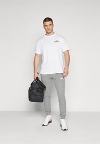 Balts T-krekls ar sarkano "ST. PAULI" logo, pelēkas sporta bikses un melna sporta soma ar melnbaltajiem akcentiem. Balti sporta apavi ar krāsainu apmalīti.
