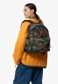 Zaino militare verde con accenti rossi, materiale testurizzato, tasca anteriore con cordino e spallacci regolabili, indossato sopra una giacca quilted arancione.