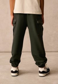 Dark green sweatpants with elastic cuffs, side pockets, and a relaxed fit, paired with black and white New Balance sneakers.