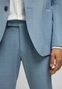 Lichtblauw pak met een gestructureerde stof, enkele knoopsluiting van de jas en op maat gemaakte broek met zijzakken, gestyled met een wit overhemd.