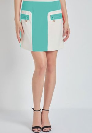 Piernas femeninas en tacones abiertos negros y una minifalda de bloques de color con un panel central en azul verdoso y paneles laterales en crema con bolsillos con cremallera.