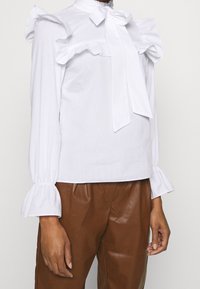 Blusa bianca con dettagli arricciati sulle spalle e sui polsini, caratterizzata da un fiocco legato al collo. Tessuto di cotone stropicciato, abbinato a pantaloni marroni.