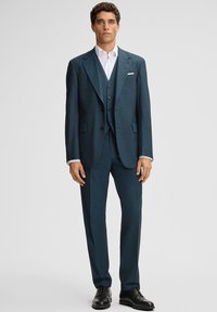 Mężczyzna w ciemnoniebieskim garniturze trzyczęściowym, z białą koszulą i czarnymi butami wizytowymi, stojący na tle jasnego, jednolitego tła.