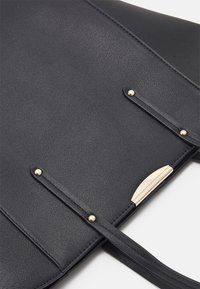 Sac à main en cuir noir avec une texture lisse, des accents en métal doré, et deux petits boutons sur les anses pour la fermeture.