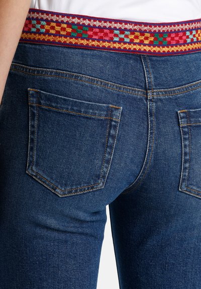 Sininen denim-housut, joissa on värikäs kudottu kangas vyötäröllä. Ominaisuuksiin kuuluu kaksi takataskua ja näkyvä ompeleminen.