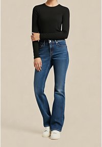 Personne portant une chemise noire à manches longues, un jean bleu taille haute et des chaussures blanches, debout sur un fond uni.