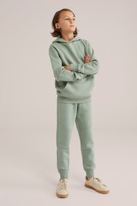 Felpa verde chiaro e pantaloni coordinati realizzati in tessuto morbido, con una vestibilità rilassata, tasche frontali e dettagli di cucitura a contrasto. Scarpe casual completano il look.