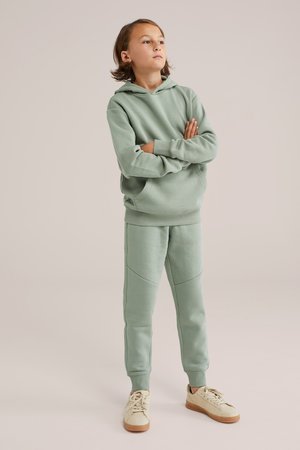 Helles grünes Hoodie und passende Hose aus weichem Stoff, mit lockerer Passform, Fronttaschen und kontrastierenden Nahtdetails. Casual-Schuhe runden das Outfit ab.