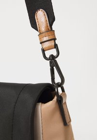 Cinghia della borsa nera e marrone con un accento in pelle marrone e chiusura in metallo nero. Tessuto testurizzato e dettagli in pelle liscia visibili.