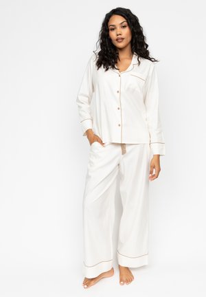 Ensemble de pyjama blanc comprenant une chemise à boutons avec une poche poitrine et des manches longues, ainsi que des pantalons amples avec des détails en piping beige contrastant.