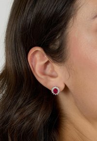 Orecchini a stud con gemma rossa incastonata in un metallo color argento, con un alone di accentuazione di diamanti, posati sull'orecchio di un modello con capelli scuri.
