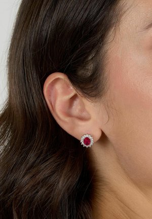 Orecchini a stud con gemma rossa incastonata in un metallo color argento, con un alone di accentuazione di diamanti, posati sull'orecchio di un modello con capelli scuri.