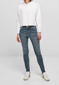 Polo blanco de manga larga recortada combinado con unos vaqueros azules de corte slim y zapatillas blancas con detalles verdes. Textura suave, estilo casual.