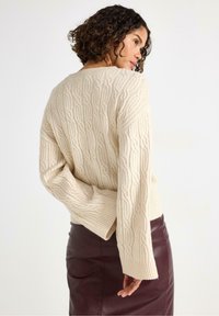 Kvinde med krøllet hår iført en cremede kabelstrik sweater og en mørkebrune læderskirt, poserende med den ene hånd bag ryggen.