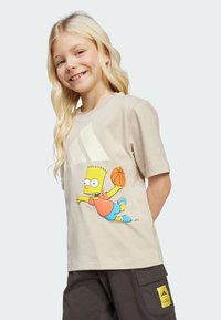Camiseta de algodón beige que presenta a un personaje de dibujos animados, Bart Simpson, sosteniendo un balón de baloncesto. Mangas cortas y corte relajado. Acentuada con un gran logo blanco.
