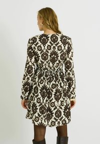Robe à manches longues avec un motif floral noir sur un fond crème, dotée d'une taille smockée et d'un ourlet évasé. Tissu lisse avec un léger éclat.