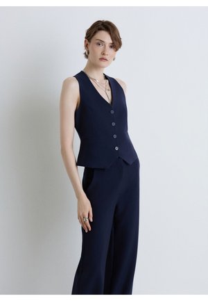 Femme aux cheveux courts portant un gilet sans manches bleu foncé boutonné et un pantalon large assorti, debout contre un mur gris clair.