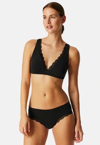 Musta alusvaatesetti, joka sisältää pitsireunaisen braletten syvällä V-kaula-aukolla ja vastaavat brief-alushousut, joissa on aaltoilevat pitsikoristeet. Pehmeää kangasta.