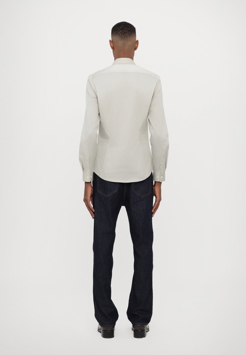 Chemise gris clair à manches longues avec un col pointu, des poignets à bouton et un ourlet arrondi, associée à un jean bleu foncé et des chaussures noires.