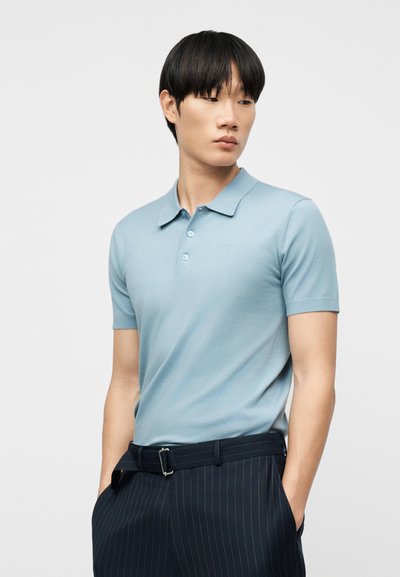 Νέος άνδρας που φοράει ανοιχτό μπλε πόλο με κοντά μανίκια και σκούρο παντελόνι με λεπτές ρίγες και ζώνη, με τα χέρια στις τσέπες, κοιτάζει προς το πλάι.